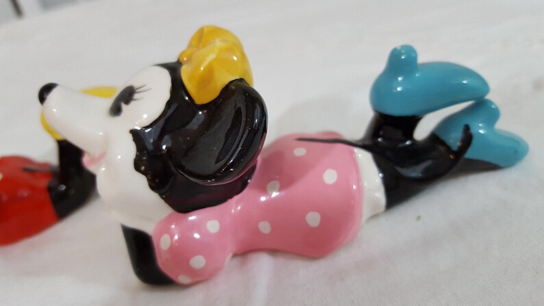 Może przedstawiać: Ceramiczna figurka Myszki Minnie w pozycji leżącej. Figurka ma białą twarz, czarne uszy i ż&oacute;łtą kokardę. Nosi r&oacute;żową sukienkę w białe grochy i niebieskie buty. Jest to przedmiot kolekcjonerski.