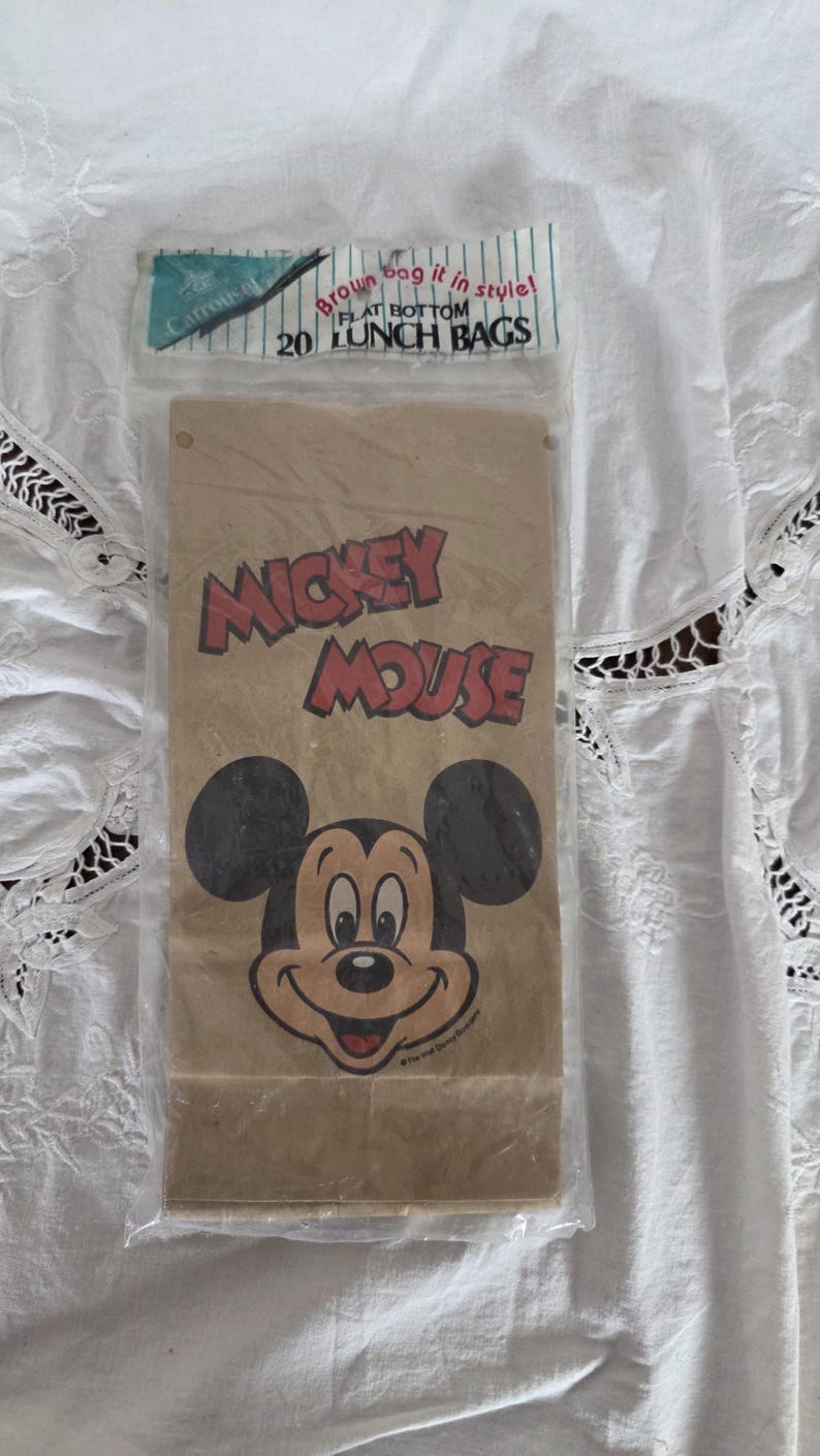 May include: A package of 20 flat-bottom lunch bags featuring a smiling Mickey Mouse graphic in red and black on a brown paper bag. The package is sealed in clear plastic.