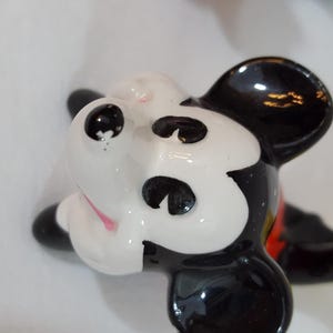 Może przedstawiać: Ceramiczna figurka Myszki Miki, z czarno-białą twarzą, czarnymi uszami, czarnymi oczami i r&oacute;żowym językiem. Figurka ma czerwoną koszulę, czarne szorty i ż&oacute;łte rękawice. Figurka leży.