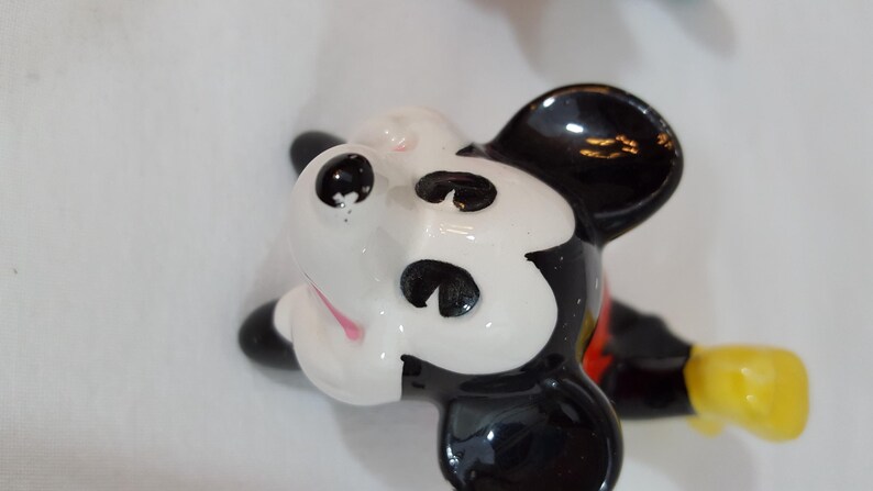 Może przedstawiać: Ceramiczna figurka Myszki Miki, z czarno-białą twarzą, czarnymi uszami, czarnymi oczami i r&oacute;żowym językiem. Figurka ma czerwoną koszulę, czarne szorty i ż&oacute;łte rękawice. Figurka leży.