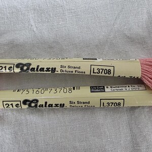 May include: Two spools of Galaxy Deluxe Floss in a soft pink hue. Each spool has a label with the text "Galaxy Six Strand Deluxe Floss L3708". The thread is neatly wound, ready for embroidery or other crafts.