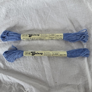 May include: Two spools of blue embroidery floss, labeled "Galaxy" and "L322 Art. 711". Each spool contains 8.7 yards of 100% double mercerized long staple cotton. The floss is a light blue color.