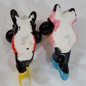 Może przedstawiać: Dwie ceramiczne figurki postaci Disneya, Myszki Miki i Myszki Minnie, widziane od tyłu. Miki ma czerwoną koszulę i ż&oacute;łte buty. Myszka Minnie ma na sobie r&oacute;żową sukienkę w białe grochy i niebieskie buty. Obie są czarno-białe.