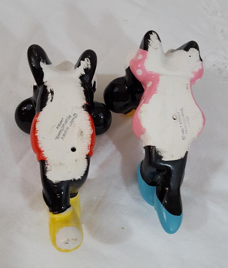 Może przedstawiać: Dwie ceramiczne figurki postaci Disneya, Myszki Miki i Myszki Minnie, widziane od tyłu. Miki ma czerwoną koszulę i ż&oacute;łte buty. Myszka Minnie ma na sobie r&oacute;żową sukienkę w białe grochy i niebieskie buty. Obie są czarno-białe.