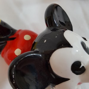Może przedstawiać: Zbliżenie ceramicznej figurki Myszki Miki. Figurka ma czarną głowę z białymi rysami twarzy i dużymi czarnymi uszami. Czerwony element w białe grochy jest widoczny w tle.