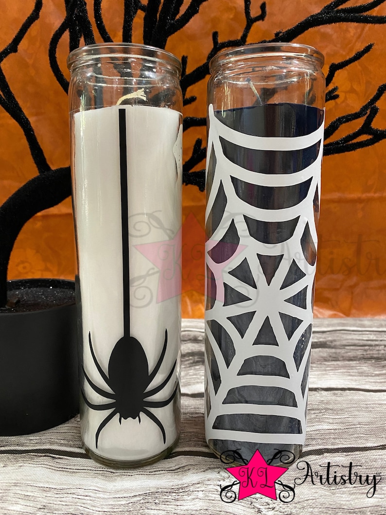 May include: Two glass candle jars with Halloween-themed designs. One jar has a black spider silhouette on a white background. The other jar has a white spiderweb design on a black background.