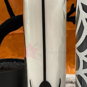 May include: A white candle in a glass jar with a black spider silhouette design. The spider is hanging from a web.