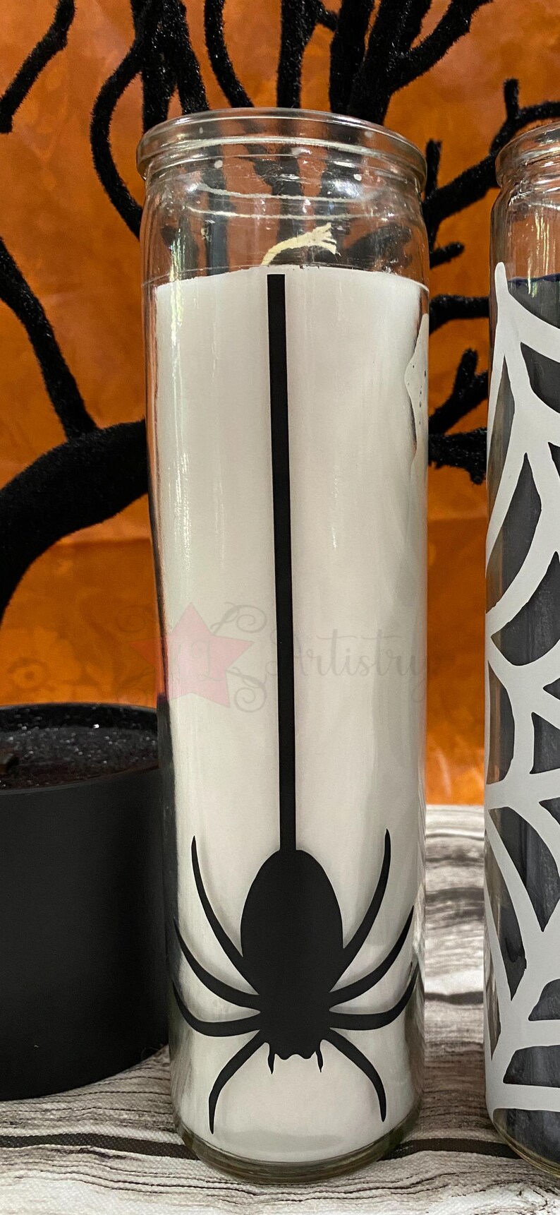May include: A white candle in a glass jar with a black spider silhouette design. The spider is hanging from a web.