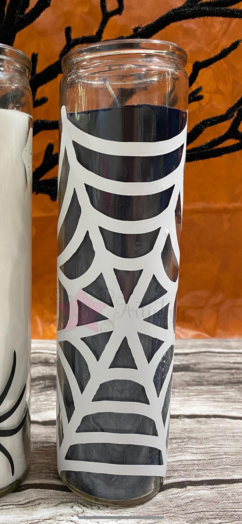 May include: A black glass candle with a white spiderweb design. The candle is in a clear glass jar.