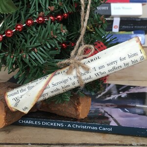 May include: A small artificial Christmas tree decorated with red beads and a paper scroll ornament with the text "CAROL ald Scrooge's no hew. I am sorry for him; I ngry with him if I ried. Who suffers by his ill meelf always. Her" and the title "CHARLES DICKENS. A Christmas Carol".