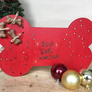 Könnte beinhalten: Roter, hölzerner Hundeknochen-förmiger Adventskalender mit dem Text "Days Until Santa Paws". Der Kalender hat Löcher zum Platzieren von Leckerlis oder anderen kleinen Gegenständen. Es befinden sich fünf Hundeknochen-förmige Leckerlis auf dem Kalender.
