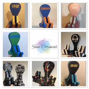 Pode incluir: Nove suportes de telefone personalizados com cores e designs diferentes. Os suportes são feitos de madeira e têm um espaço para um telefone e um suporte para auscultadores. Os suportes são decorados com cores e padrões diferentes, incluindo preto, azul, verde, rosa e branco. Os suportes são personalizados com nomes, incluindo Cruz, Cameron, Lara, Fletcher, Max, Jude, Jack, Oliver, e um suporte com um design de pintura salpicada.