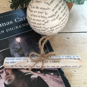 May include: A Christmas bauble made from a book page with the text "A Christmas Carol" printed on it. The bauble is tied with twine and is hanging from a Christmas tree.