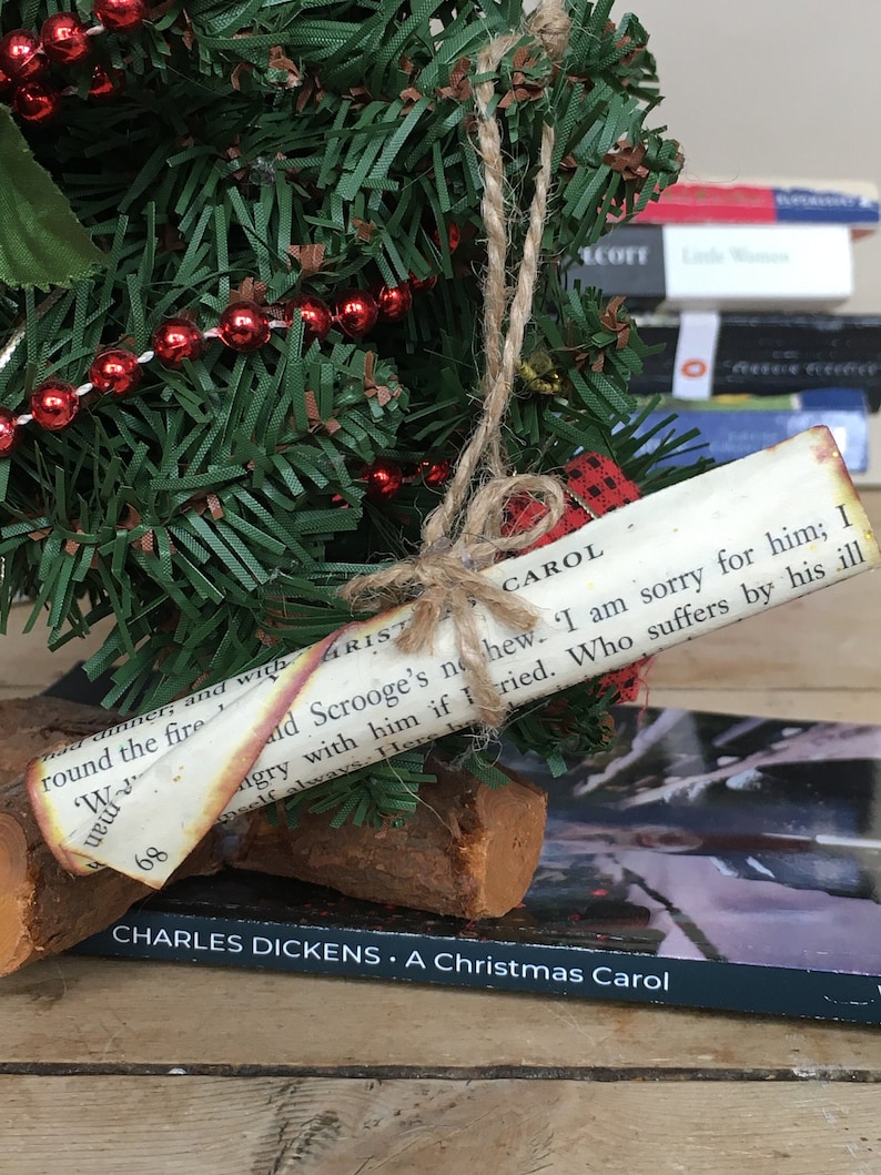 May include: A small artificial Christmas tree decorated with red beads and a paper scroll ornament with the text "CAROL ald Scrooge's no hew. I am sorry for him; I ngry with him if I ried. Who suffers by his ill meelf always. Her" and the title "CHARLES DICKENS. A Christmas Carol".