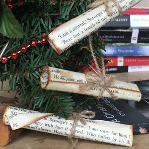 May include: Three vintage-style paper scrolls with text from "A Christmas Carol" tied with twine and hanging from a small artificial Christmas tree with red beads. The scrolls are on a wooden surface.