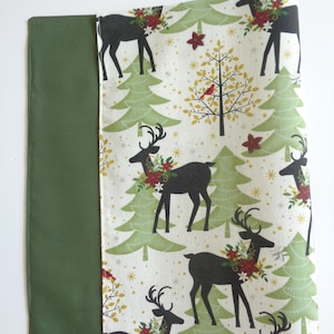 Puede incluir: Un camino de mesa con temática navideña con un borde verde y un centro estampado blanco. El patrón incluye ciervos negros con coronas florales, árboles de Navidad verdes y flores de pascua rojas. El camino de mesa es de tela.