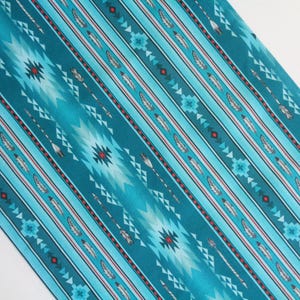 May include: Teal fabric with a Native American-inspired design. The pattern includes white arrows, feathers, and geometric shapes, with red and light blue accents. The fabric is diagonally oriented.