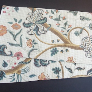 May include: Two rectangular placemats with a cream-colored background and a floral pattern. The design features brown branches, teal and blue leaves, and orange and pink flowers. The placemats are made of a textured fabric.