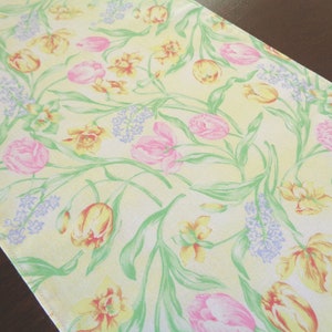 May include: A floral table runner with a pattern of pink and yellow tulips, daffodils, and purple hyacinths on a pale yellow background. Green stems and leaves intertwine throughout the design. The runner is made of fabric.