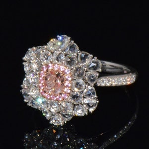 Puede incluir: Un anillo de oro blanco con una piedra central de diamante rosa y blanco rodeada de diamantes blancos m&aacute;s peque&ntilde;os. El anillo tiene una banda con peque&ntilde;os diamantes blancos.