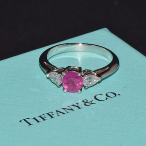 May include: A pink sapphire engagement ring with two diamond accents set in a platinum band. The ring is displayed in a Tiffany & Co. blue box.