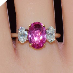 May include: A three-stone ring featuring a large oval-cut vibrant pink center gemstone flanked by two smaller oval-cut clear diamonds. The stones are set in a yellow gold band. A white diagonal banner in the corner displays the text GIA.