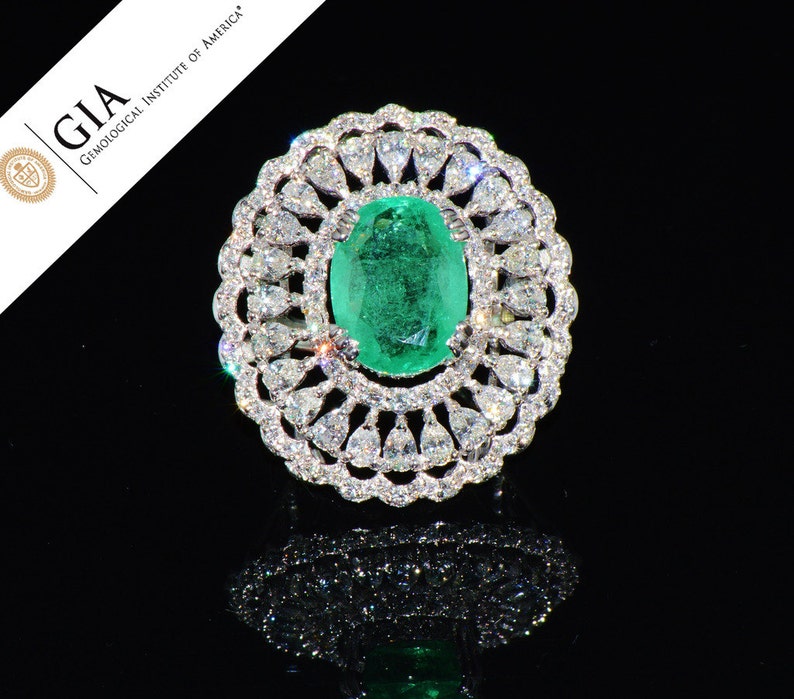 May include: A ring featuring a large, oval emerald gemstone encircled by a halo of smaller diamonds. The ring is set in white gold and boasts a delicate, intricate design.