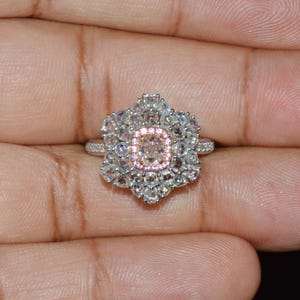 Puede incluir: Un anillo de plata con una gran piedra preciosa rosa de forma ovalada rodeada de piedras preciosas blancas m&aacute;s peque&ntilde;as. El anillo est&aacute; engastado en un dise&ntilde;o floral.