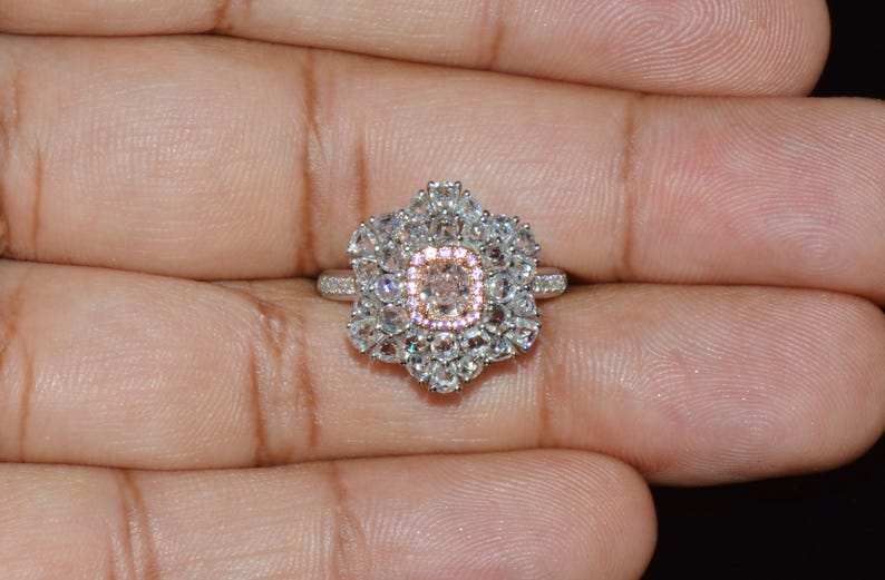 Puede incluir: Un anillo de plata con una gran piedra preciosa rosa de forma ovalada rodeada de piedras preciosas blancas m&aacute;s peque&ntilde;as. El anillo est&aacute; engastado en un dise&ntilde;o floral.