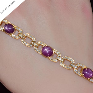 May include: A gold bracelet with three red star sapphires and diamond accents. The bracelet is on a person's wrist.