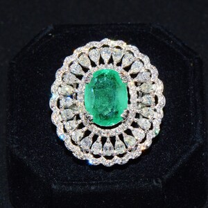 May include: A silver ring featuring a large oval emerald gemstone surrounded by smaller diamonds. The ring is set in a delicate floral design.