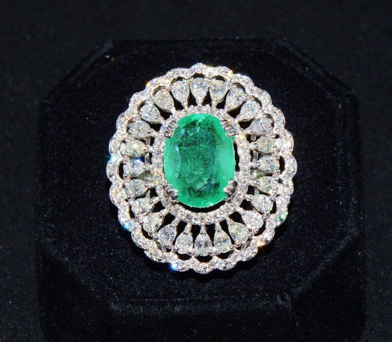 May include: A silver ring featuring a large oval emerald gemstone surrounded by smaller diamonds. The ring is set in a delicate floral design.