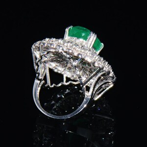 May include: A silver ring featuring a large emerald green gemstone surrounded by smaller diamonds. The ring is set in a delicate, intricate design.