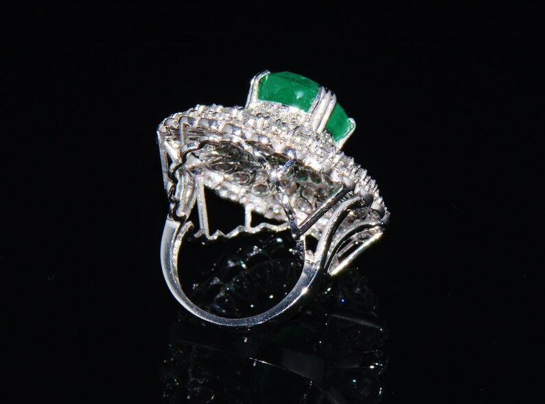 May include: A silver ring featuring a large emerald green gemstone surrounded by smaller diamonds. The ring is set in a delicate, intricate design.