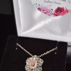 Puede incluir: Un collar de cadena de plata con un colgante de piedra preciosa rosa y blanca. El colgante tiene forma de flor con una piedra preciosa rosa grande en el centro rodeada de piedras preciosas blancas m&aacute;s peque&ntilde;as.