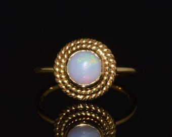May include: A gold ring with a round, opalescent gemstone. The gemstone is surrounded by a twisted rope design. The ring is set against a black background, with a reflection of the ring visible.