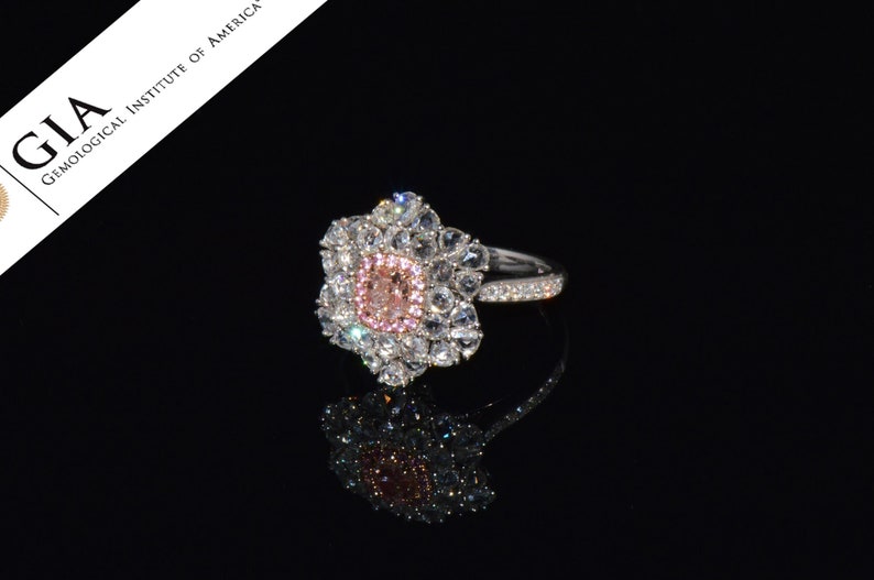 Puede incluir: Un anillo de oro blanco con una piedra central de diamante rosa y blanco rodeada de diamantes blancos m&aacute;s peque&ntilde;os. El anillo tiene una banda con peque&ntilde;os diamantes blancos.