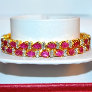 May include: A gold bracelet with pink pear-shaped gemstones and smaller round white gemstones. The bracelet is displayed in a red and white box.
