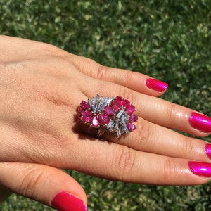 May include: A ring featuring a large cluster of pink gemstones surrounded by smaller diamonds. The ring is set in gold and is worn on a finger with pink nail polish.