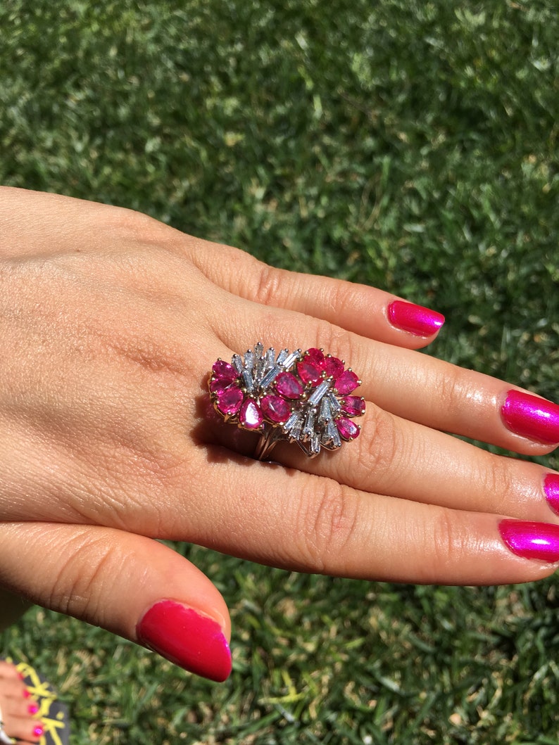 May include: A ring featuring a large cluster of pink gemstones surrounded by smaller diamonds. The ring is set in gold and is worn on a finger with pink nail polish.