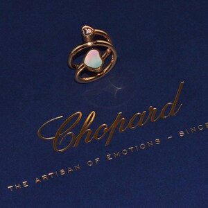 May include: A gold-toned ring featuring a double-band design with a small heart-shaped accent at the top and a larger, iridescent heart-shaped element in the center. The ring is placed on a dark blue surface with the gold text Chopard THE ARTISAN OF EMOTIONS - SINCE 1860.