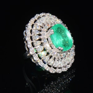 May include: A silver ring featuring a large, oval emerald gemstone surrounded by smaller diamonds. The ring has a delicate, intricate design.