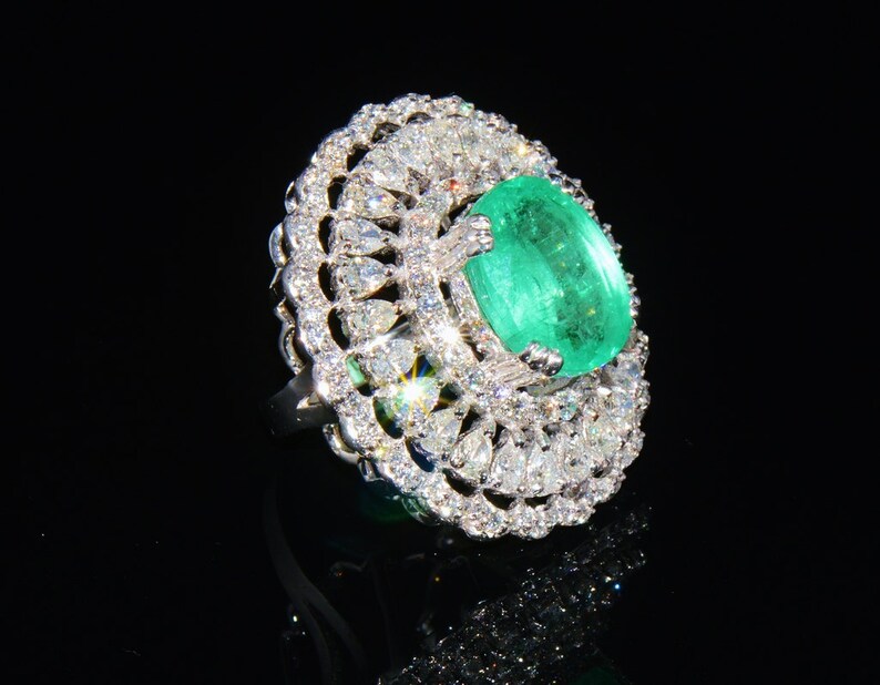 May include: A silver ring featuring a large, oval emerald gemstone surrounded by smaller diamonds. The ring has a delicate, intricate design.