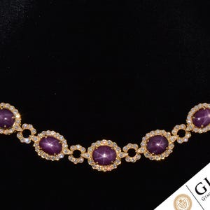 May include: A gold necklace with oval, deep purple gemstones and small, clear gemstones. The necklace has an intricate design against a black backdrop. The GIA logo is visible in the bottom right corner.