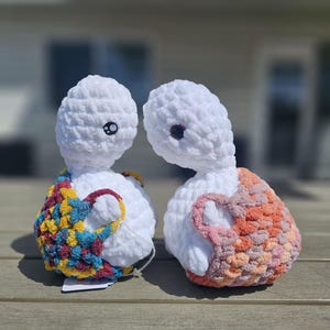 May include: Two white crocheted plush figures with round heads and dark eyes. The left figure wears a multicolored backpack with blue, yellow, and red yarn. The right figure wears a peach and pink crocheted backpack. Both have soft textures.