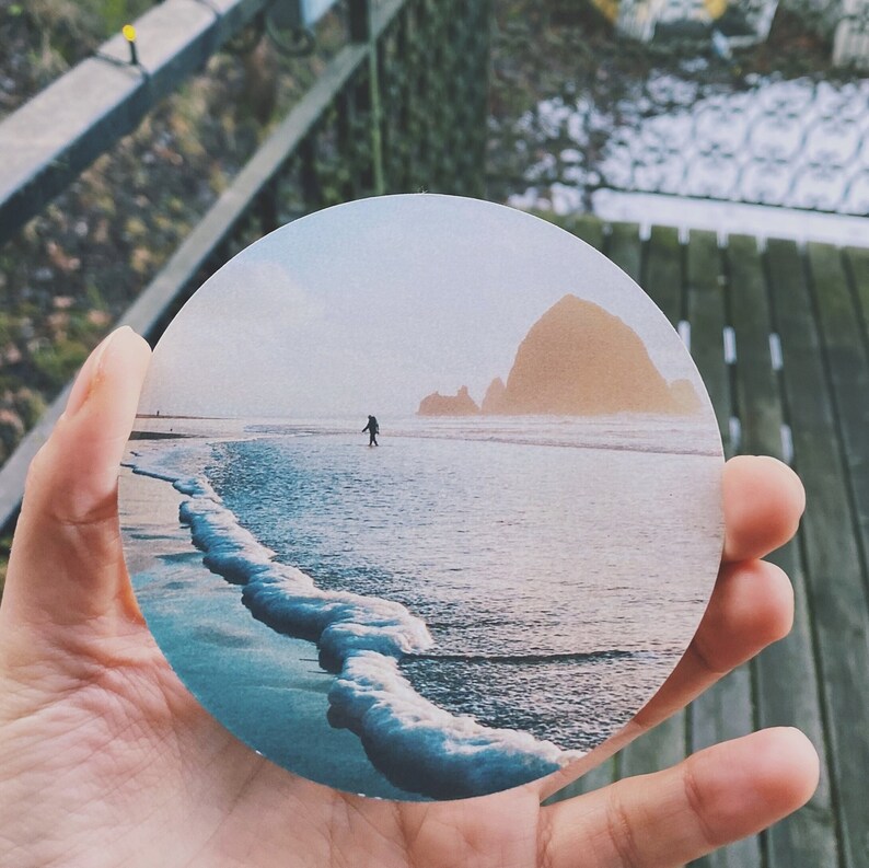 May include: A round coaster featuring a scenic photograph of a beach with a large rock formation in the distance. The beach is sandy with blue water and white waves crashing on the shore. A lone figure walks along the beach.