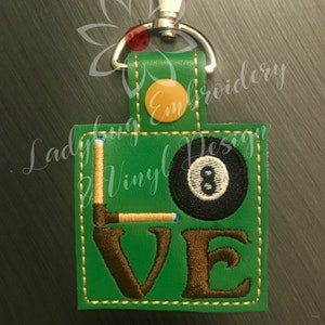 Könnte beinhalten: Grüner Schlüsselanhänger mit dem Wort "LOVE", das aus einem Billardqueue und einer Achtkugel gebildet wird. Er hat einen silbernen Verschluss und einen orangefarbenen Knopf. Der Text "Ladybug Vinyl Design" ist ebenfalls sichtbar.