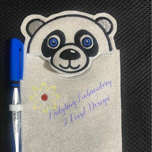 Puede incluir: Un portalápices de oso panda gris brillante con un bolígrafo azul. El panda tiene detalles en blanco y negro con ojos azules. El soporte tiene una margarita y el texto "Ladybug Embroidery & Vinyl Design".