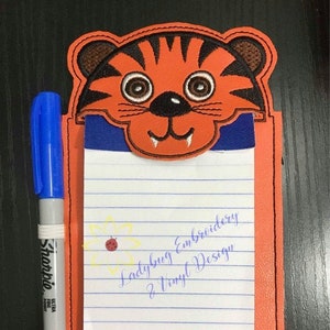 May include: Orange notepad with a tiger face design at the top. The notepad has lined paper and the text "Ladybug Embroidery & Vinyl Design". A blue Sharpie marker is on the left side.