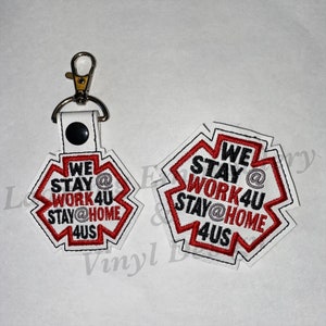 May include: Two white and red embroidered patches with the text "WE STAY @ WORK4U STAY@HOME 4US". One patch is attached to a keychain with a silver clasp and a black snap. The patches have a black border.